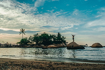 Man standing on rock with arms raised at tropical beach sunset