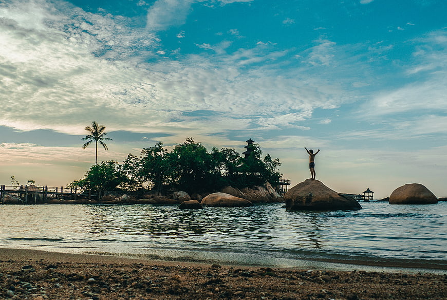 Man standing on rock with arms raised at tropical beach sunset