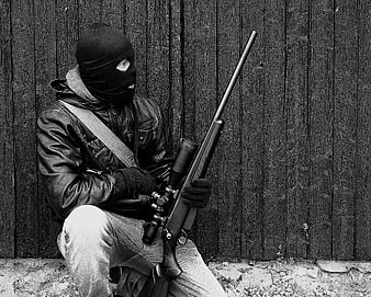 Masked sniper holding rifle against dark wooden fence in black and white
