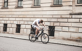 Man Cycling on City Street Past Stone Building