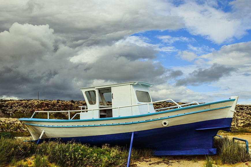 White and Blue Fishing Boat on Rocky Shore Under Cloudy Sky