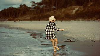 Child in plaid shirt and hat running on wet beach