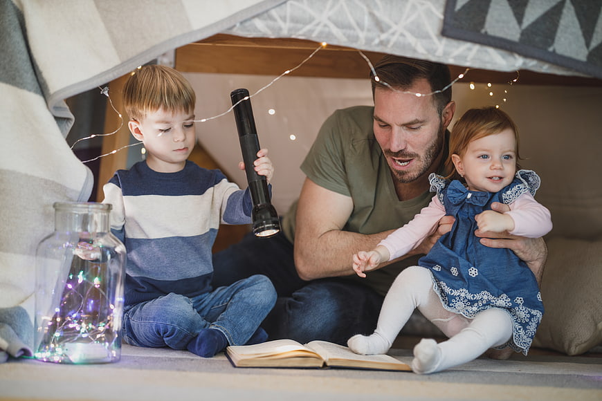 Father reading to children in cozy blanket fort with fairy lights