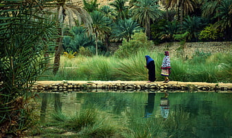 Two women in traditional dress walking by a pond in an Omani garden