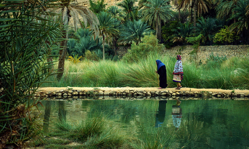Two women in traditional dress walking by a pond in an Omani garden