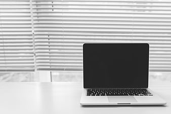 Silver laptop with black screen on white desk near window blinds