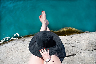 Woman relaxing on rocks by turquoise sea with black hat