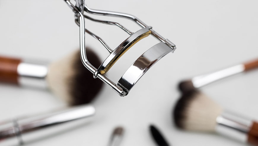 Close-up of silver eyelash curler with makeup brushes