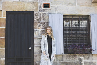 Blonde woman in long coat standing by rustic stone house