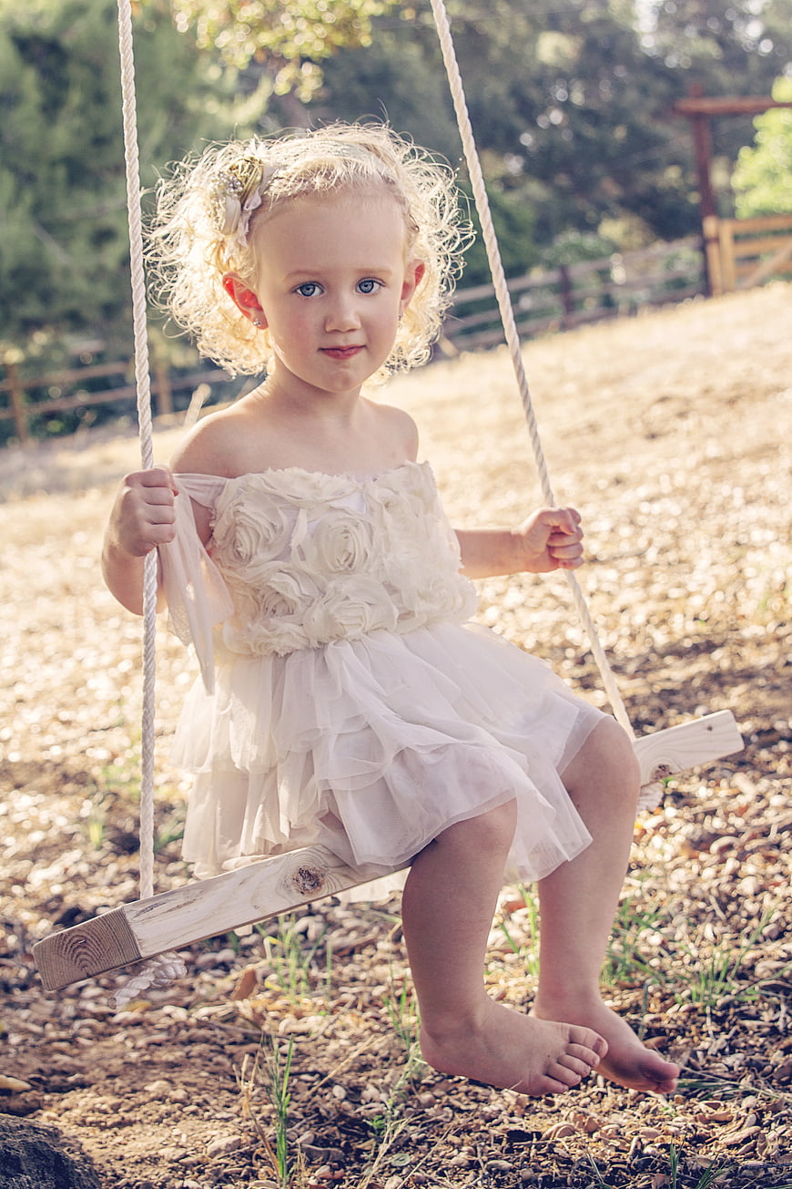 Cute blonde toddler girl in white dress sitting on wooden swing outdoors