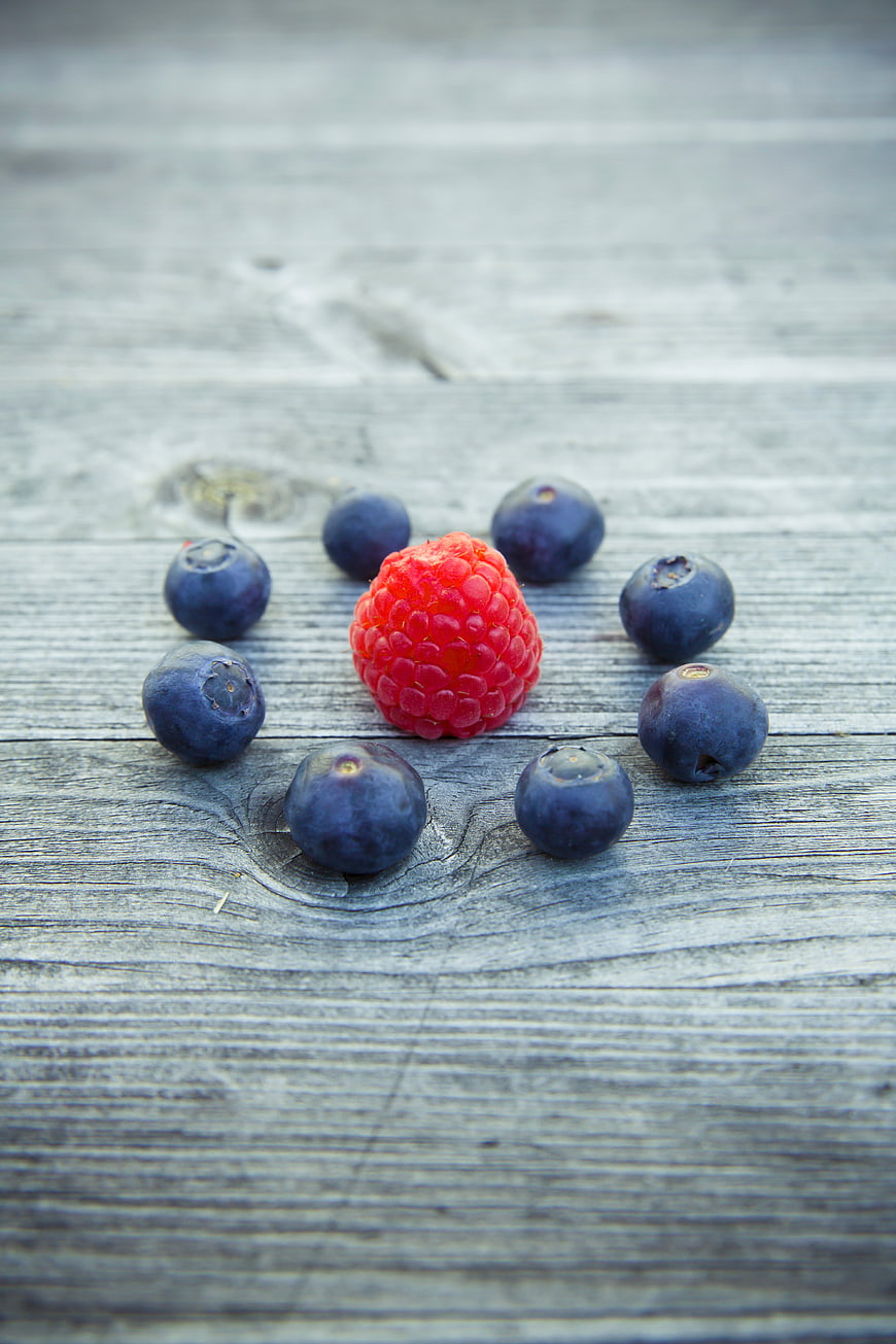 Single Raspberry Surrounded by Blueberries on Weathered Wood