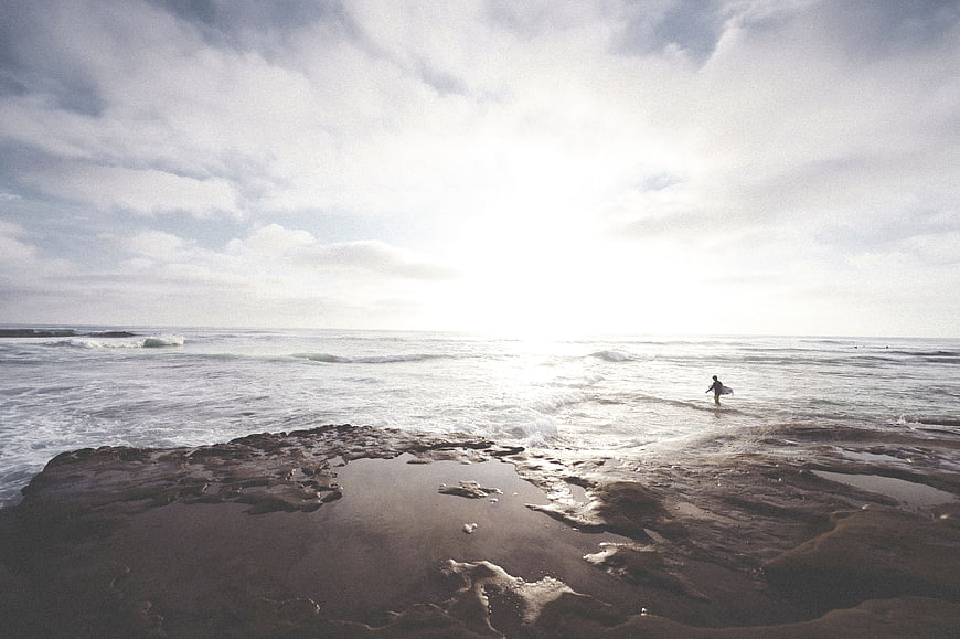 Lone Surfer Standing in Shallow Ocean Water on Rocky Shore