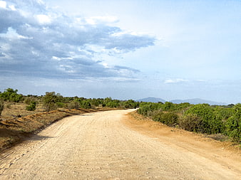 Empty dirt road winding through rural landscape with green bushes and cloudy sky
