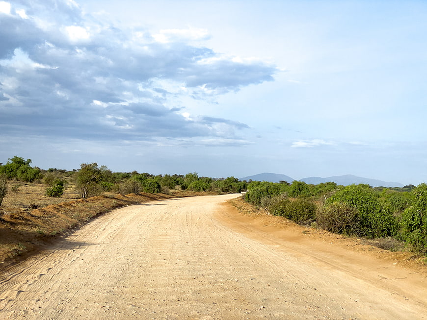 Empty dirt road winding through rural landscape with green bushes and cloudy sky