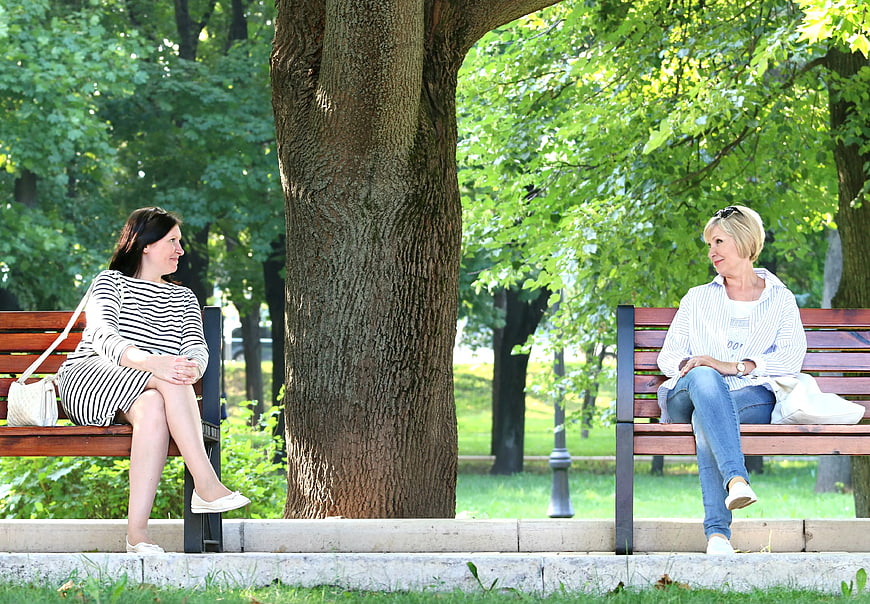 Two women chatting on separate park benches under a large tree