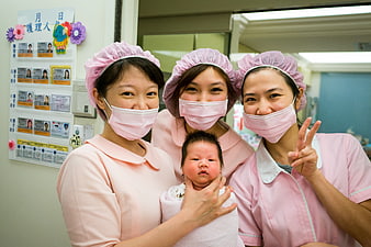 Three nurses in pink uniforms holding a newborn baby in a hospital