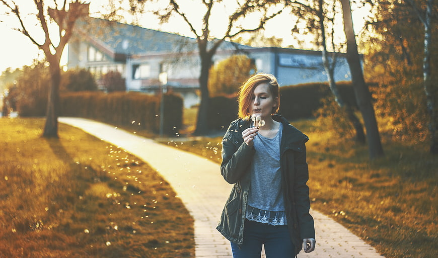 Young Woman Blowing Dandelion Seeds in Autumn Park