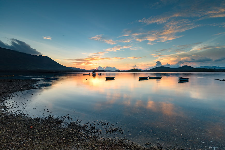 Serene Sunrise Over Calm Sea With Boats And Mountain Reflections
