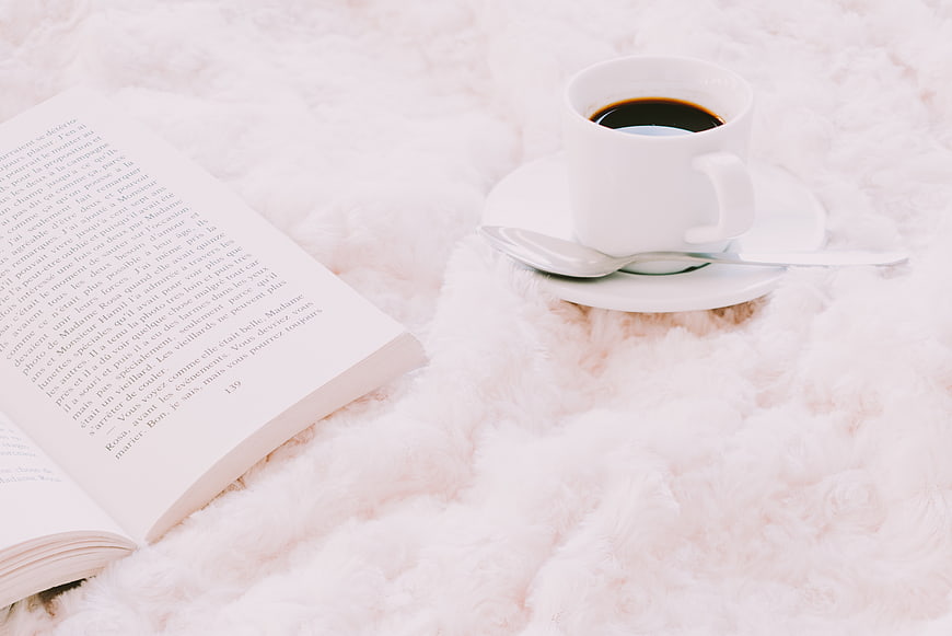 Cozy coffee cup and open book on fluffy white blanket