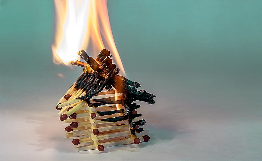 Burning matchstick house structure with flames on teal background