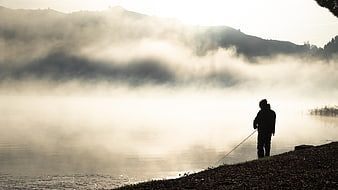 Silhouette of fisherman on misty lake shore at dawn