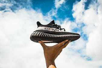 Hand holding black sneaker against blue sky with clouds