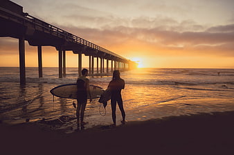 Surfers with boards standing on beach at sunset near pier