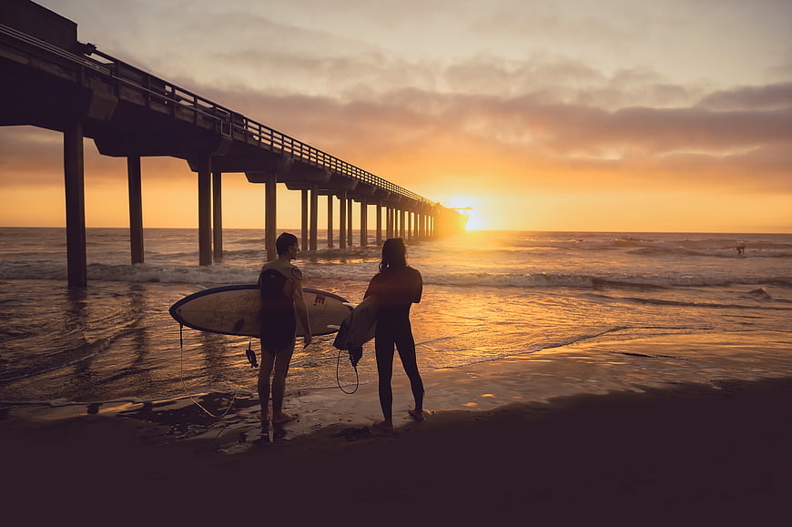 Surfers with boards standing on beach at sunset near pier