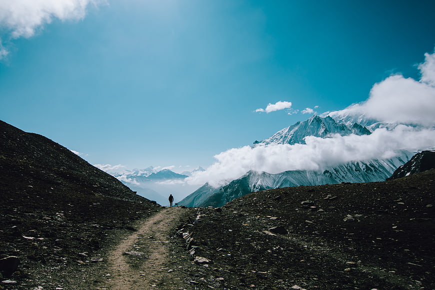Lone Hiker on Mountain Trail with Snowy Peaks and Clouds