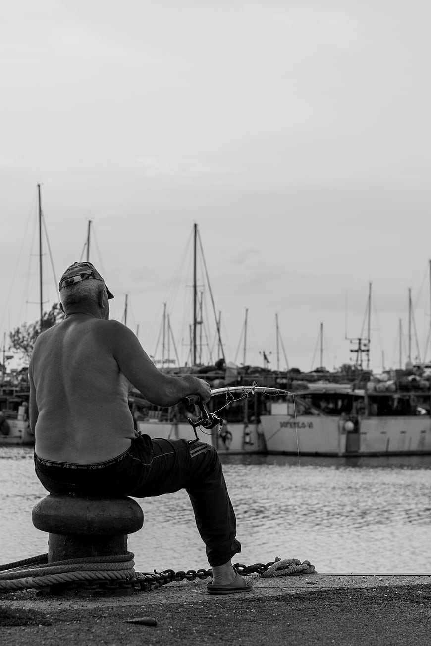 Shirtless man fishing on a harbor bollard in black and white