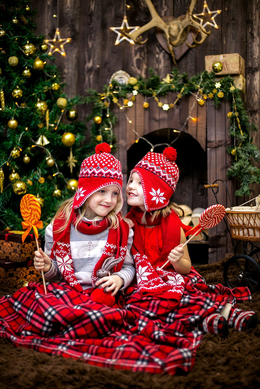 Two girls in red winter hats laughing with lollipops by Christmas tree