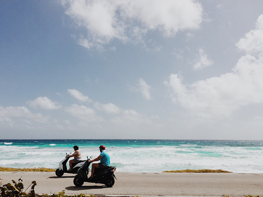 Two Men Riding Scooters Along Coastal Road by Ocean
