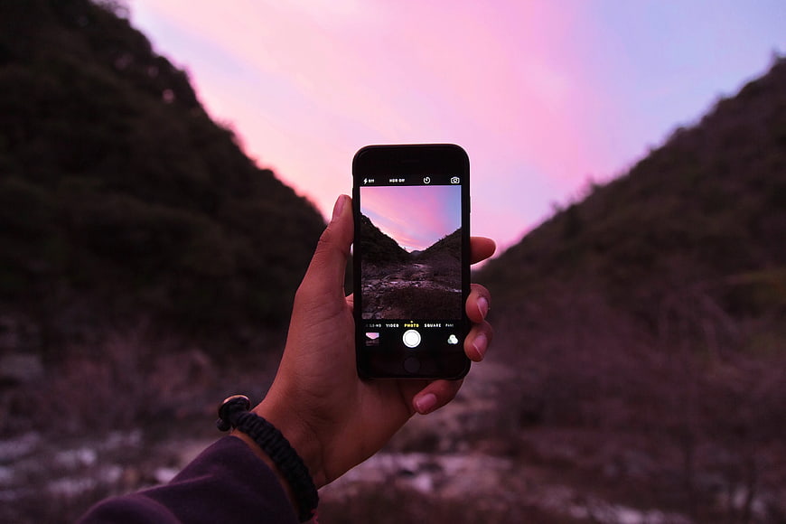 Hand holding smartphone capturing pink sunset valley landscape