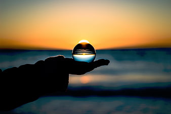 Hand holding crystal lens sphere capturing sunset over ocean horizon
