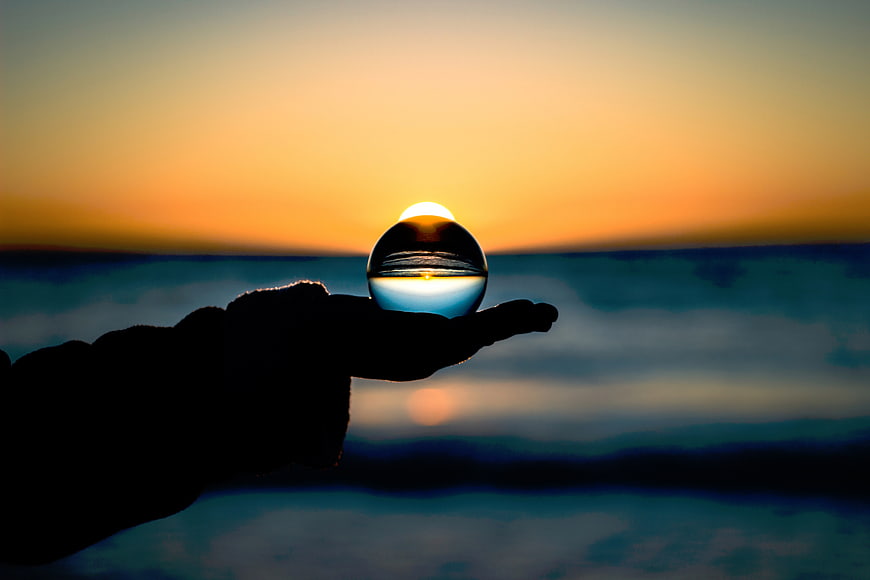 Hand holding crystal lens sphere capturing sunset over ocean horizon
