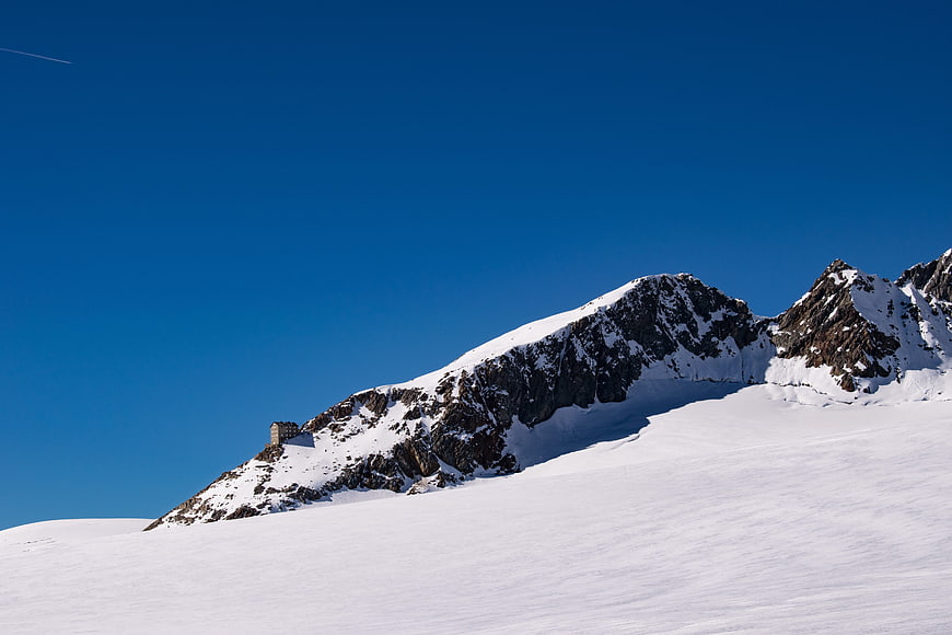 Snowy mountain peak with small building against blue sky