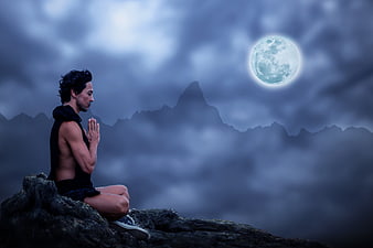 Man meditating on rocky cliff under full moon at night