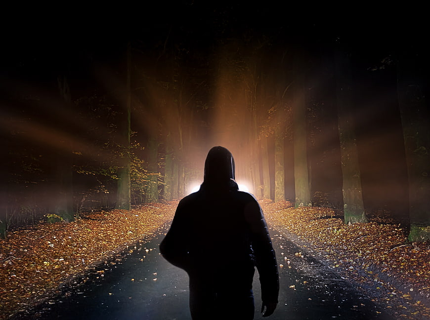 Silhouette of hooded figure walking on dark forest path at night