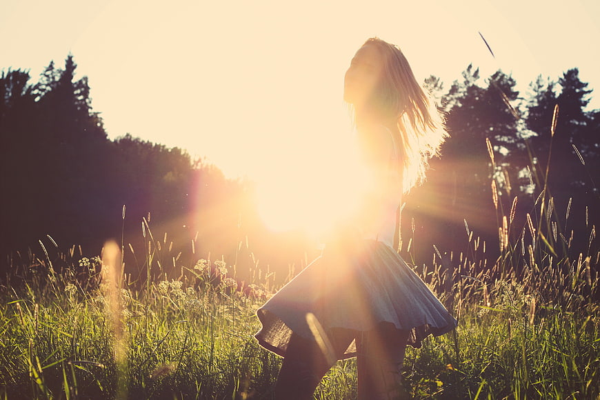 Woman in white dress standing in grassy field with sunset lens flare