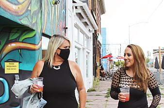 Two women walking on city street with drinks and face mask