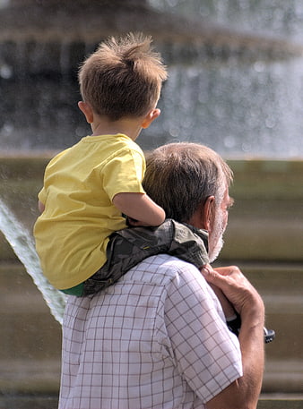 Grandfather Carrying Grandson On Shoulders At Outdoor Fountain