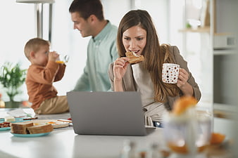 Mother eating breakfast while working on laptop with family