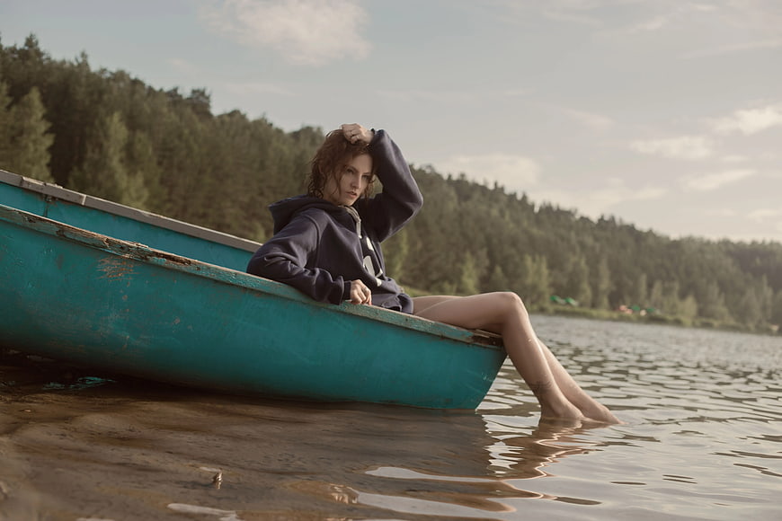 Woman in hoodie sitting in teal canoe by lake shore