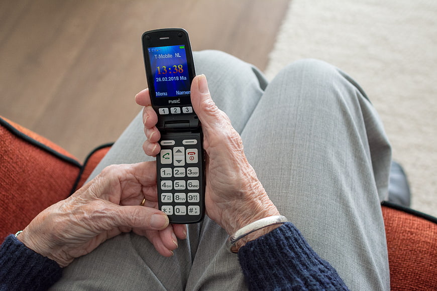 Elderly Woman Holding Black Flip Phone On Lap Indoors