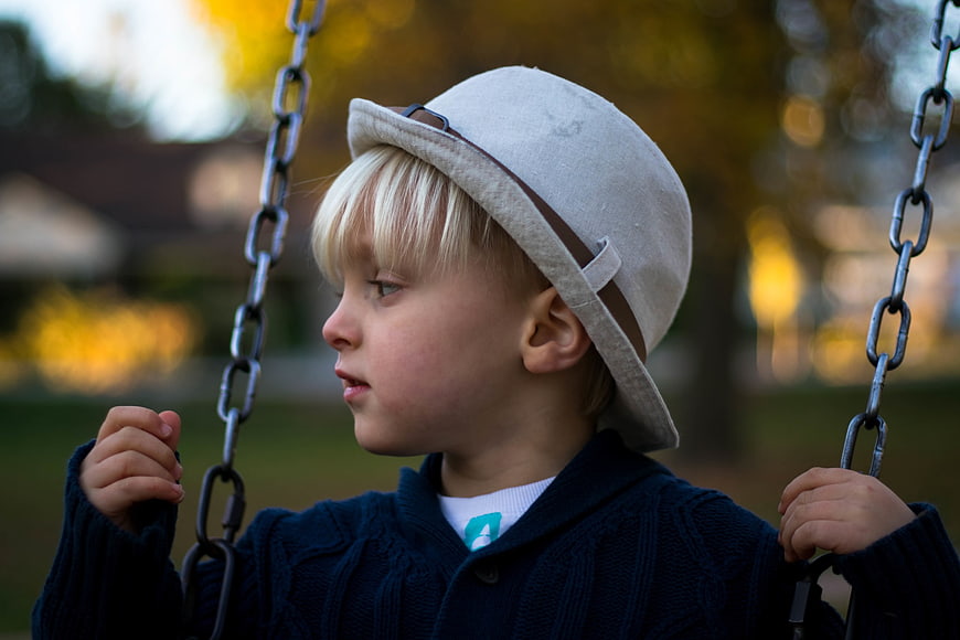 Cute Blonde Boy in Fedora Hat Sitting on Playground Swing