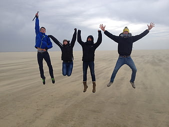 Four friends jumping joyfully on a vast sandy beach under an overcast sky