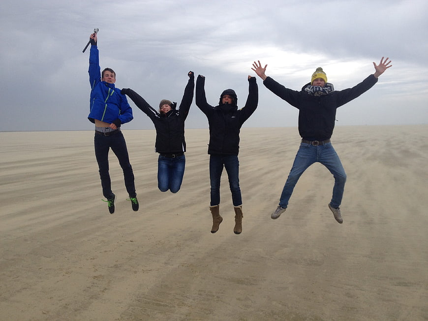 Four friends jumping joyfully on a vast sandy beach under an overcast sky