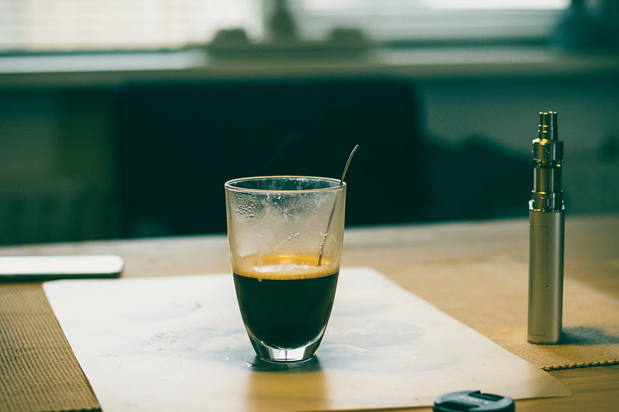 Half-empty espresso glass and vape pen on wooden table
