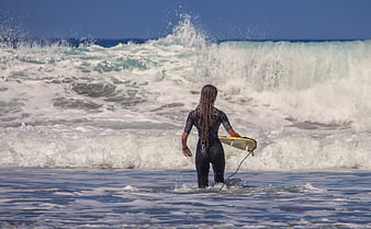 Female surfer in wetsuit walking into ocean waves with surfboard