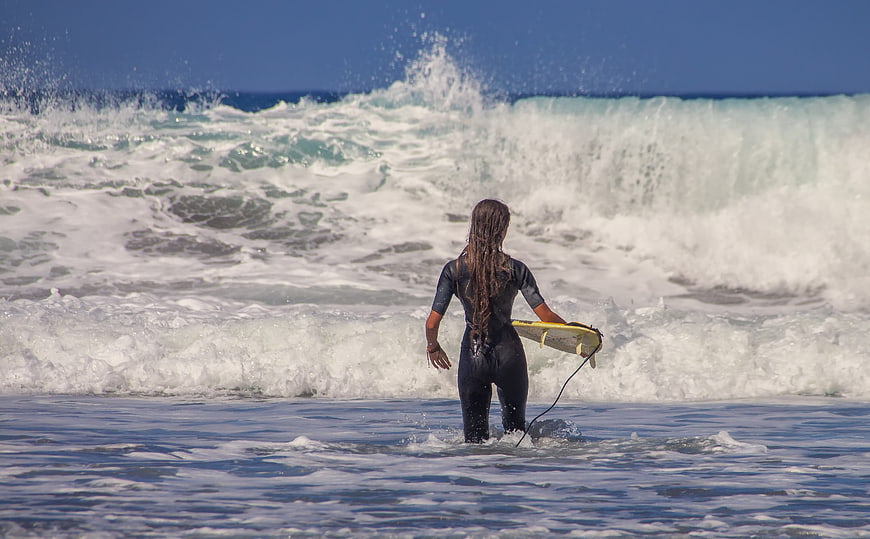 Female surfer in wetsuit walking into ocean waves with surfboard